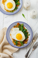 Fried egg with potato pancake, arugula and avocado on ceramic plate for breakfast on white wooden table background. Selective focus. Top view. Copy space.