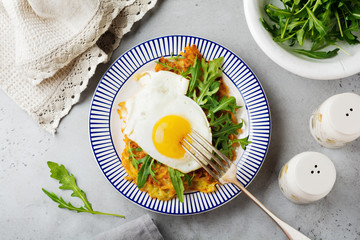 Fried egg with potato pancake, arugula and avocado on ceramic plate for breakfast on gray old concrete background. Selective focus. Top view. Copy space.