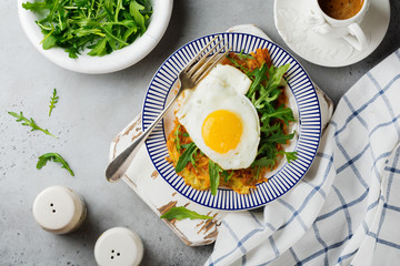 Fried egg with potato pancake, arugula and avocado on ceramic plate for breakfast on gray old concrete background. Selective focus. Top view. Copy space.
