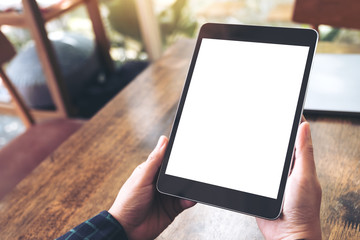 Mockup image of hands holding black tablet pc with blank desktop white screen on wooden table in...