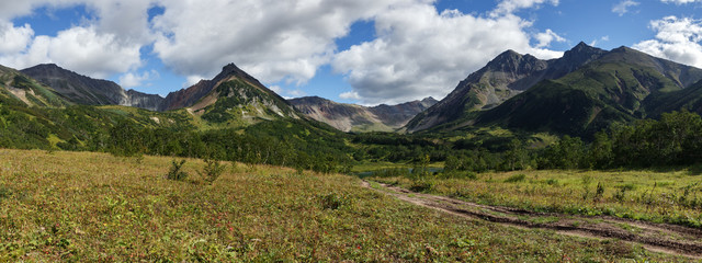 Beautiful panorama mountain landscape of Kamchatka Peninsula: early autumn view of Mountain Range Vachkazhets with forest slopes of hills, glade grass covered and clouds in blue sky on sunny day.