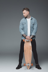 full length view of stylish young tattooed man standing with skateboard on grey