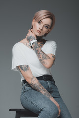 beautiful tattooed girl in white t-shirt sitting on stool and looking at camera isolated on grey