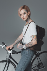 Fototapeta na wymiar beautiful tattooed girl with backpack standing with bicycle and looking at camera isolated on grey