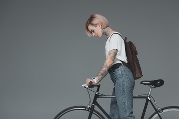 Fototapeta na wymiar side view of young woman with backpack standing with bicycle isolated on grey