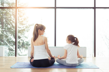 Obraz premium Young beautiful woman and charming little daughter are doing difficult yoga exercises and smiling while working out at gym with beautiful scenery outside window with sunlight. Healthy fitness concept