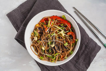 Healthy vegan buckwheat noodles with a mix of vegetables, sprouts and nut-soy sauce. Vegetarian padthai soba noodles in white bowl on gray background. Top view