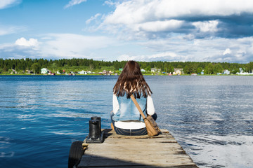 Young woman in jeans clothes sitting on mooring near river and looking on shore