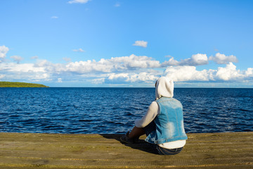 Lonely young woman sitting on wooden mooring near lake and looking on nature, relaxation concept