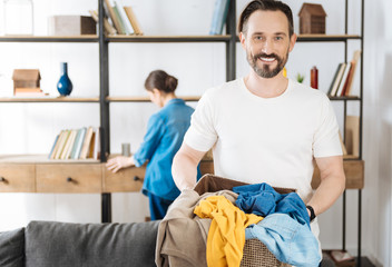 Time for laundry. Bearded confident jolly husband posing on the blurred background while smiling to the camera and carrying basket