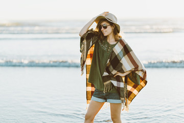Charming young girl with poncho, hat, sunglasses with cute smile standing in water on the beach at sunset in warm weather. Boohoo style.
