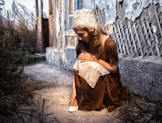 sad woman in a rustic dress sitting near old brick wall in old house and trying to dress a white...
