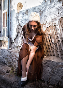 sad woman in a rustic dress sitting near old brick wall in old house and trying to dress a white shoe. Cinderella style