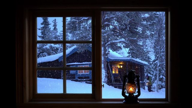 Time lapse of woman carrying a lantern walking by a mountain cabin in wintry conditions seen through a window.