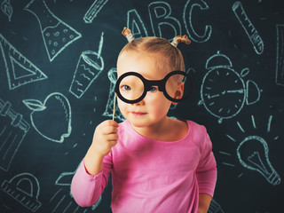 Pretty small girl investigate something through glasses on chalkboard background, back to school theme