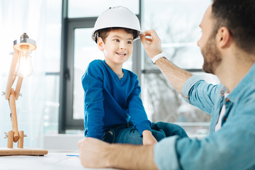 Loving parent. Young loving father fixing a white hard hat on the head of his little son sitting on the top of the table in his office and chatting with him