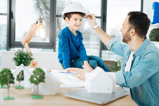 Gentle care. Pleasant loving father adjusting a hard hat on the head of his little son sitting on the table in fathers office while the man working on a blueprint