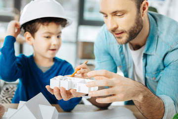 Thorough examination. Handsome young father and his little son in a white hard hat sitting at the table and examining a 3D house layout model