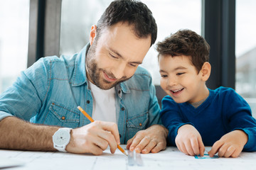 Under supervision. Upbeat little boy sitting at the table next to his father and watching him draw a blueprint with a pencil and a ruler