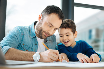 Interested glances. Cute chubby boy sitting at the desk next to his father and watching him draw a...