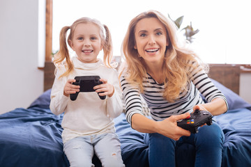 Playing with mum. Adorable content fair-haired girl laughing and holding a remote control playing with her mom while sitting on the bed