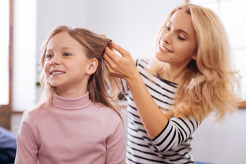 My daughter. Beautiful happy blond young mother smiling and making a hairdo for her little daughter while standing behind her child
