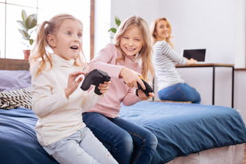 Game time. Lovely vigorous fair-haired little sisters smiling and holding remote controls while playing and hair mother working on the laptop