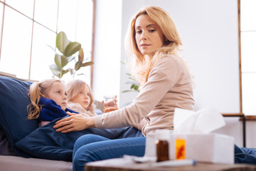 Feeling bad. Serious troubled fair-haired mother sitting near her children while her sick daughters lying in bed and looking at the medications lying on the table