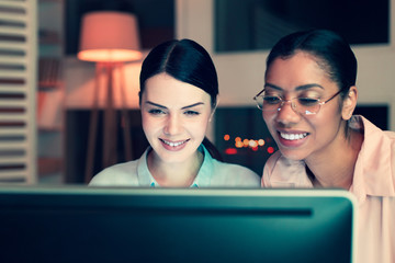 Nice pastime. Two upbeat young women watching a video on the computer together and smiling while having a break at work