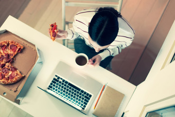Good at multitasking. The top view of a dark-haired woman in a striped pullover sitting at the table in the office and working while eating pizza and drinking tea