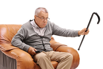 Annoyed senior with a cane sitting in an armchair and watching television