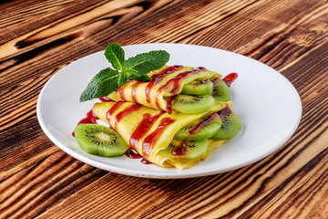 On a light table, a white plate with sliced kiwi fruits wrapped in pancakes. Pancakes are poured with jam and decorated with green mint leaves.