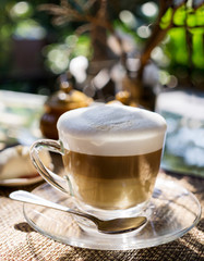 Layered cappuccino in glass cup on table at Thai cafe in warm sunlight outdoor. Blurred nature in background. Travel concept. Text space