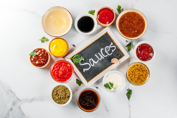 Set of different sauces - ketchup, mayonnaise, barbecue, soy, teriyaki, mustard, grain hills, pesto, adzhika, chutney, tkemali, pomegranate sauce on white marble background. Top view copy space