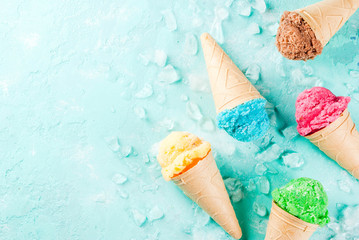 Selection of various bright multicolored ice-cream in ice cream cones - chocolate vanilla blueberry strawberry pistachio orange, on light blue sunny background, copy space top view