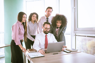 Fototapeta premium Teamwork process. Team lead sitting at the table and showing chart on laptop to his employees, who stay behind him.