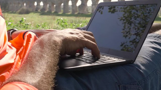 Cyclist with bike sitting on a rock under a tree writing on laptop in front of ancient roman aqueduct. Young attractive athletic man with orange sportswear and backpack in parco degli acquedotti in