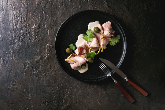 Vitello tonnato italian dish. Thin sliced veal with tuna sauce, capers and coriander served on black plate with fork and knife over dark texture background. Top view, copy space