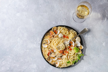 Pasta spaghetti Vongole in tomato cream sauce in black plate with fork and glass of white wine over gray texture background. Top view, copy space