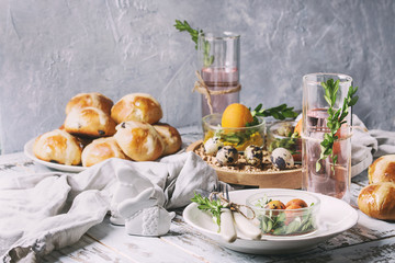 Easter table setting with colored orange eggs, hot cross buns, green branches decorated, empty white plate with cutlery, glass of lemonade drink over white plank wooden table with textile tablecloth.