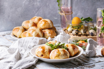 Easter table setting with colored orange eggs, plate of hot cross buns, green branches decorated, glass of cocktail drink over white plank wooden table with textile tablecloth.