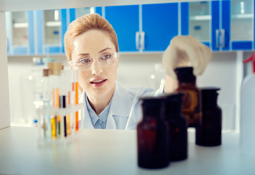 Let me see. Selective focus on a mature lady wearing safety glasses standing in a lab and taking a dark brown bottle with a chemical substance.