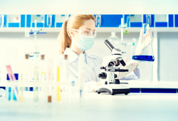 Fototapeta premium Attentive analysis. Selective focus on a mature woman standing at a microscope and examining a flask with a navy blue chemical liquid.