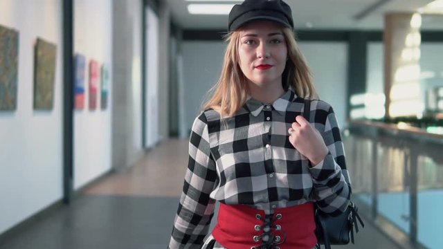 Young stylish woman wears black hat walks with bag on her shoulder in modern building, indoor slowmotion