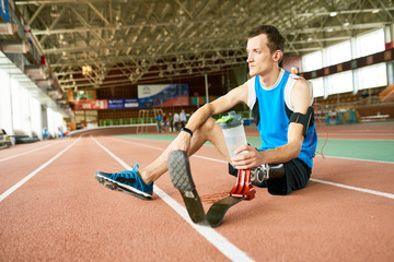 Portrait of pensive amputee athlete sitting on running track taking break from practice  to relax and drink water, copy space