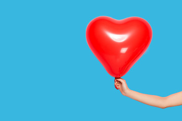 Female hand holding a red ball in the shape of a heart, isolated on a blue background.