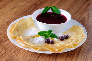 Pancakes are sprinkled with powdered sugar and decorated with green mint leaves and few currants. In a plate, a cup of jam.