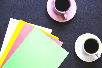 Stylish office table desk, flat lay, top view. Workplace with two cups of coffee and stack of colored papers, black concrete background, copy space