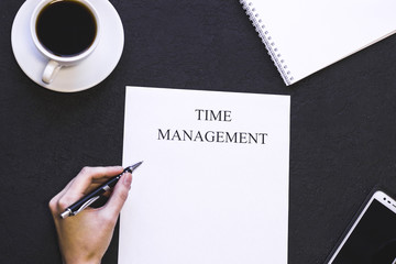 Stylish office table desk, flat lay,top view. Workplace with a coffee cup, smartphone and a paper with printed Time Management inscription, man's hand is writing, black concrete background, copy space