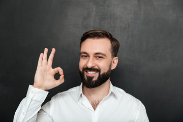 Close up photo of bearded guy smiling and gesturing with OK sign expressing good choice, being isolated over graphite background